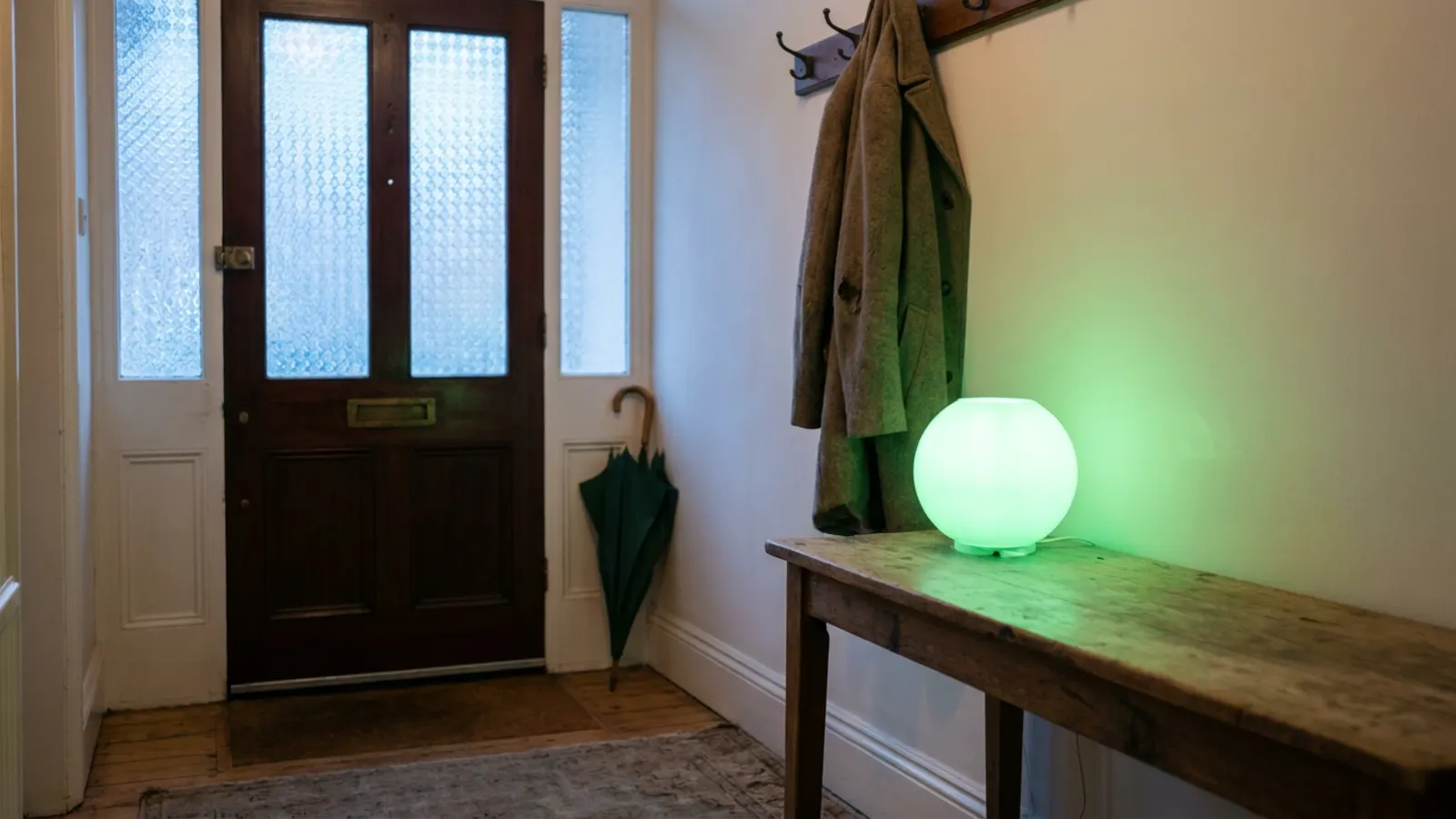 A hallway at dawn with a globe lamp glowing green on a side table, a coat on the hook and an umbrella by the door